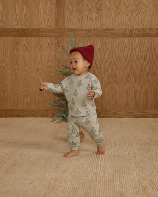 Rylee & Cru Long Sleeve Tee + Pant Set - Trees
Child wearing a red hat and patterned pajamas holding a small tree against a wooden wall.