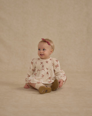 Rylee & Cru Sophia Top + Bloomer Set - Ruby Bows
Baby sitting on a beige background wearing a white shirt with brown patterns and brown shoes.