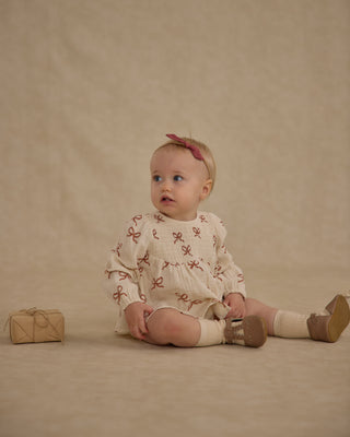 Rylee & Cru Sophia Top + Bloomer Set - Ruby Bows
Baby sitting on a beige background wearing a white outfit with red patterns and brown shoes.