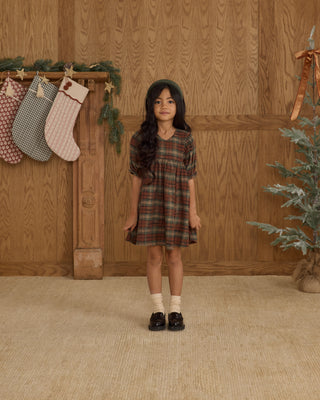 Rylee & Cru Jolene Dress - Holiday Plaid
Young girl in a plaid dress standing in front of a wooden wall with stockings and a Christmas tree.