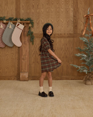 Rylee & Cru Jolene Dress - Holiday Plaid
Young girl in a plaid dress standing in front of a wooden wall with stockings and a small tree.