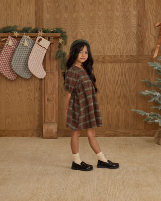 Rylee & Cru Jolene Dress - Holiday Plaid
Young girl in a plaid dress standing in front of a wooden wall with stockings and a Christmas tree.