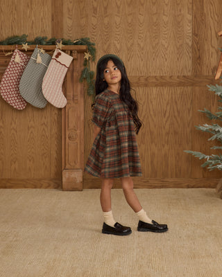 Rylee & Cru Jolene Dress - Holiday Plaid
Young girl in a plaid dress standing in front of wooden paneling with stockings hanging.