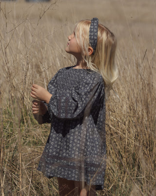 Rylee & Cru Selene Dress - Heart Ditsy
A young girl with blonde hair stands in a field of dry grass, wearing a loose, short-sleeved dark gray patterned dress and a matching patterned headband.