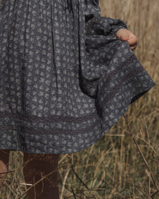 Rylee & Cru Selene Dress - Heart Ditsy
A close-up of a person outdoors wearing a dark patterned dress, holding a small brown bag, with dry grass in the background.