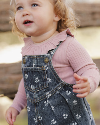 Rylee & Cru Ruffle Collar Bodysuit - Rosewood
A toddler girl wearing a dusty pink ribbed long-sleeve top with a ruffled collar and dark blue denim overall dress with a white floral print, photographed outdoors.