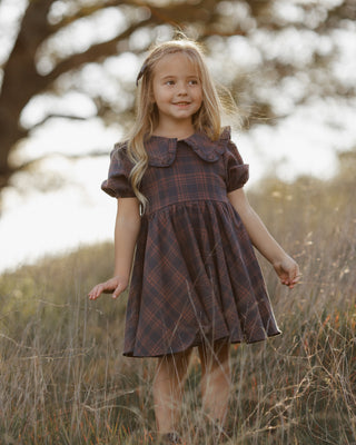 Rylee & Cru Camille Dress - Midnight Plaid
A young girl with long blonde hair stands in a field wearing a dark brown and red plaid dress with a ruffled Peter Pan collar.