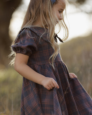 Rylee & Cru Camille Dress - Midnight Plaid
A young girl with long blonde hair wearing a dark blue and red plaid dress, standing outdoors in a softly blurred natural setting.