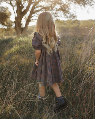 Rylee & Cru Camille Dress - Midnight Plaid
A young girl with long blonde hair stands in a grassy outdoor field, viewed from behind. She is wearing a dark plaid dress with puff sleeves and dark socks with boots.