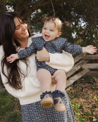 Rylee & Cru Fern Romper - Heart Ditsy
A woman outdoors holds a smiling toddler wearing a navy and white floral long-sleeve romper with a large ruffled collar, a cream knit sweater, and tan shoes.