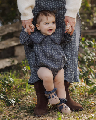 Rylee & Cru Fern Romper - Heart Ditsy
A baby wearing a dark blue and gray floral print romper, held up by an adult wearing brown boots and a cream sweater in an outdoor garden setting.