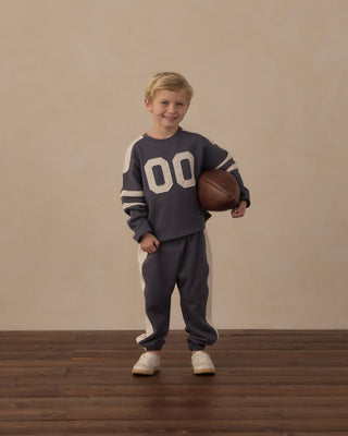 Play by Rylee & Cru Hockey Sweatshirt - Midnight
Play by Rylee & Cru Paneled Sweatpant - Midnight
A young boy stands on a wooden floor against a beige wall, wearing a dark gray athletic-style sweatshirt and sweatpants set with cream stripes and a large cream '00' on the front, while holding a brown basketball.