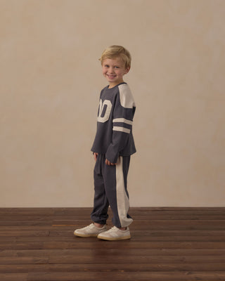 Play by Rylee & Cru Paneled Sweatpant - Midnight
A young boy stands on a dark wood floor against a warm beige wall, wearing a matching dark gray and cream athletic lounge set with a large number 0 on the chest.