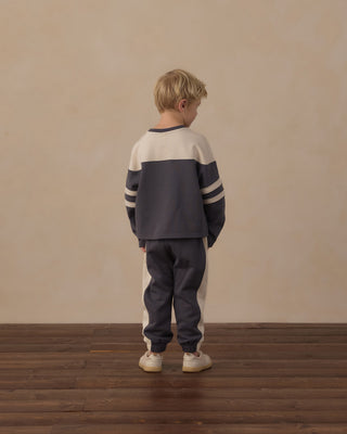 Play by Rylee & Cru Hockey Sweatshirt - Midnight
A young boy with blonde hair stands with his back to the camera, wearing a color-blocked sweatshirt and sweatpants set in navy blue and cream, standing on a dark wood floor against a textured beige wall.