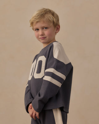 Play by Rylee & Cru Hockey Sweatshirt - Midnight
A young blond boy wearing a dark gray and cream athletic jersey with the number 00 on the front, posed against a solid beige studio backdrop.