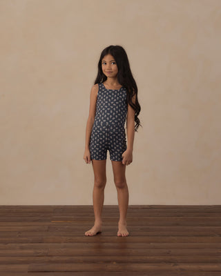 Play by Rylee & Cru Malibu Bodysuit - Blue Blooms
A young girl with long dark hair stands barefoot on a wooden floor against a textured beige wall, wearing a sleeveless dark blue romper with a small white pattern.