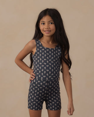 Play by Rylee & Cru Malibu Bodysuit - Blue Blooms
A young girl with long dark hair wearing a navy blue sleeveless romper with a white floral pattern, posing against a solid beige studio background.