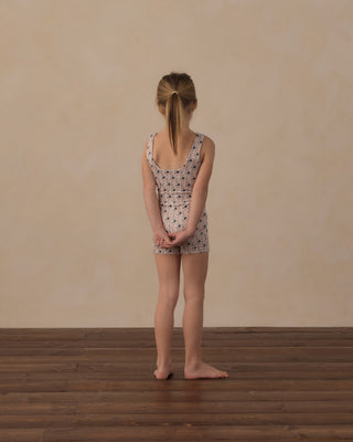 Play by Rylee & Cru Malibu Bodysuit - Blue Primrose
A young girl with blonde hair in a ponytail stands with her back to the camera, wearing a sleeveless one-piece romper with a small navy blue pattern on a light beige background. She is standing on a dark wood floor against a textured beige wall.