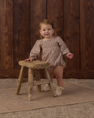 Rylee & Cru Adelaide Romper - Primrose
A toddler girl wearing a long-sleeve beige romper with a brown floral pattern, standing next to a rustic wooden stool in front of a dark wood paneled wall.