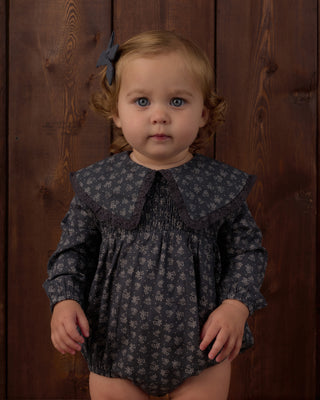 Rylee & Cru Fern Romper - Heart Ditsy
A toddler girl with light brown hair and blue eyes wearing a dark gray floral print romper with a wide smocked collar, standing against a dark wood plank background.