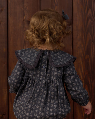 Rylee & Cru Fern Romper - Heart Ditsy
A child viewed from behind wearing a dark gray long-sleeve blouse with a small white floral pattern, a large ruffled collar, and a dark bow in their hair, standing against a dark wood plank background.