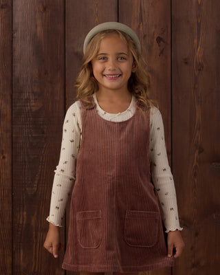 Rylee & Cru Odette Overall Dress - Mulberry
A young girl smiling in front of a dark wood plank wall, wearing a brown corduroy pinafore dress over a cream long-sleeve shirt with small dark motifs, a cream beret, and a small cream bag.