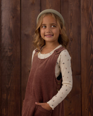 Rylee & Cru Ribbed Long Sleeve - Natural Ditsy
Rylee & Cru Odette Overall Dress - Mulberry
A young girl with blonde hair wearing a beige beret, a cream long-sleeve shirt with small dark floral prints, and a brown corduroy pinafore overalls, standing against a dark wood plank background.