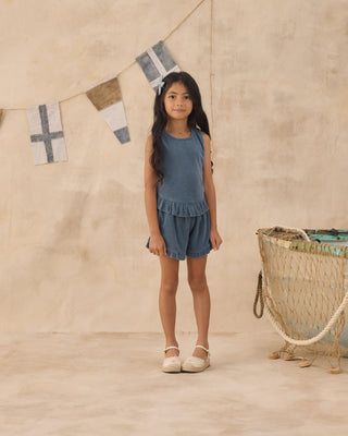 Rylee & Cru Faye Set - Marine
Young girl in a blue outfit standing in front of a beige wall with decorative flags and a basket.