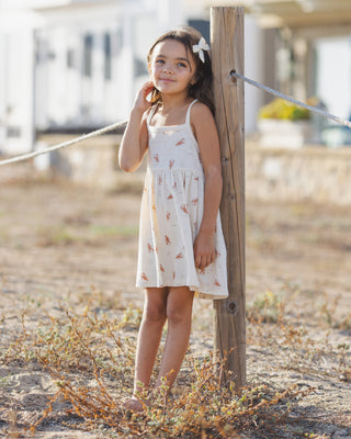 Rylee & Cru Lea Dress - Lobster
Young girl in a white dress with floral patterns standing next to a wooden post on a beach.