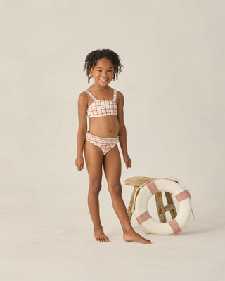 Rylee & Cru Rouched Bikini - Cherry Check
Young girl in a checkered bikini standing next to a life buoy on a beige background