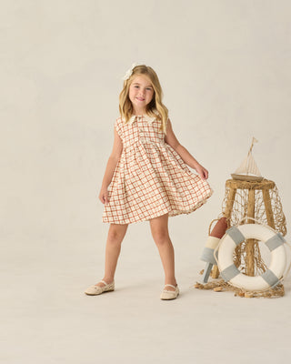 Rylee & Cru Collared Babydoll Dress - Cherry Check
Young girl in a checkered dress standing next to a life buoy and wooden stool on a plain background