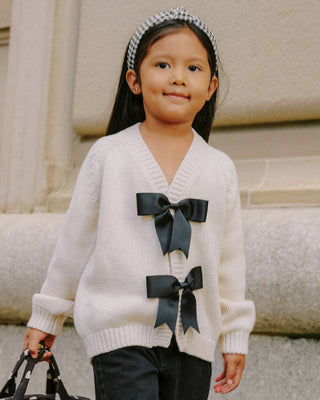 Rylee & Cru Bow Cardigan - Natural
Child wearing a white sweater with black bows in front of a stone building.