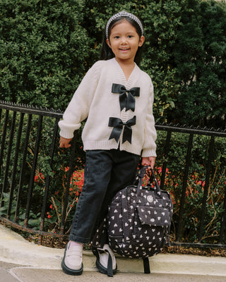 Rylee & Cru Bow Cardigan - Natural
Child wearing a white sweater with black bows, standing next to a black and white patterned suitcase.