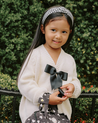 Rylee & Cru Bow Cardigan - Natural
Young girl holding a black bow with a white sweater and checkered headband, standing against a green hedge.