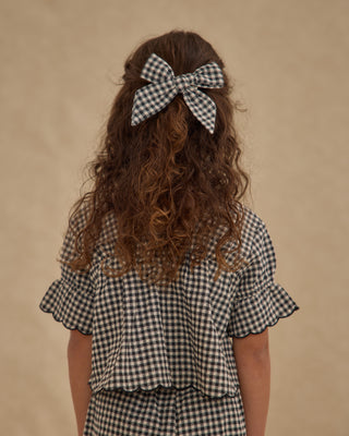 Rylee & Cru Bow - Black Gingham
Person wearing a checkered shirt with a large bow in their hair on a beige background