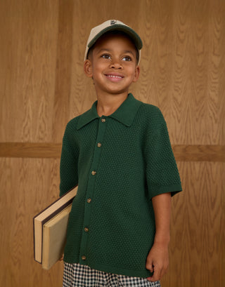 Rylee & Cru Aiden Polo - Green
Child wearing a green shirt and cap, holding books against a wooden paneled background
