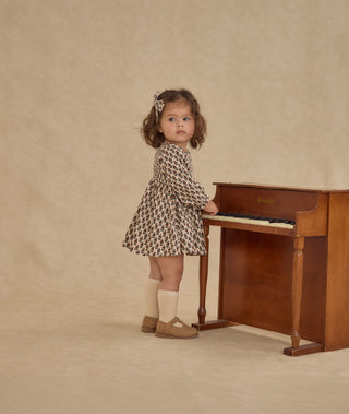 Rylee & Cru Capri Dress - Harvest Floral
Young girl in a patterned dress standing next to a wooden piano against a beige background