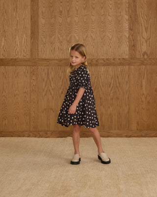 Rylee & Cru Rose Dress - Hearts
Young girl wearing a black dress with white patterns against a wooden paneled wall.