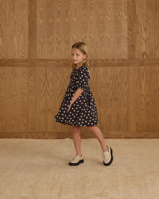 Rylee & Cru Rose Dress - Hearts
Young girl in a black dress with white patterns standing against a wooden paneled wall.