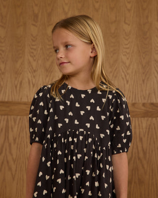 Rylee & Cru Rose Dress - Hearts
Young girl wearing a black dress with white patterns against a wooden paneled wall.