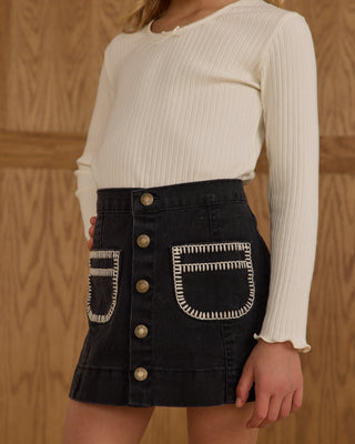 Rylee & Cru Denver Mini Skirt -  Washed Black
Person wearing a white ribbed sweater and black denim skirt with gold buttons and embroidery against a wooden background.