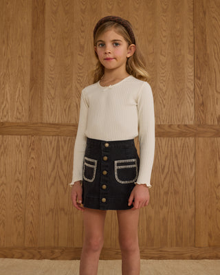 Rylee & Cru Denver Mini Skirt -  Washed Black
Young girl wearing a white long-sleeve top and black skirt with button details against a wooden paneled wall.