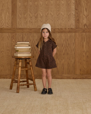 Rylee & Cru Utility Romper - Leopard
Young girl in a brown dress standing next to a wooden stool with books against a wooden paneled wall.
