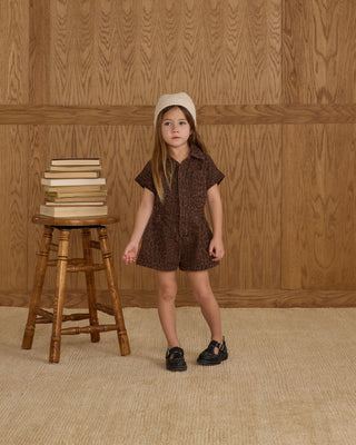 Rylee & Cru Utility Romper - Leopard
Young girl in a brown dress standing next to a wooden stool with books against a wooden paneled wall.