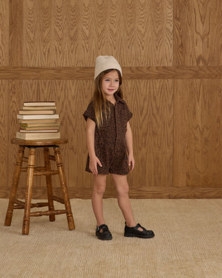 Rylee & Cru Utility Romper - Leopard
Young girl in a brown patterned dress and black shoes standing next to a wooden stool with books against a wooden paneled wall.