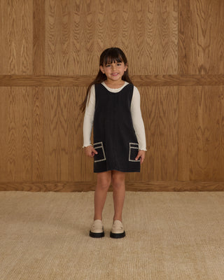 Rylee & Cru V-Neck Overall Dress - Washed Black
Young girl wearing a black dress with white sleeves against a wooden paneled wall.