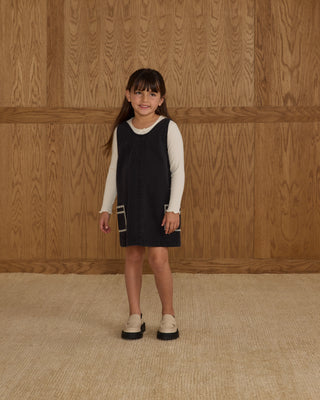 Rylee & Cru V-Neck Overall Dress - Washed Black
Young girl wearing a black dress with a white shirt underneath against a wooden paneled wall.
