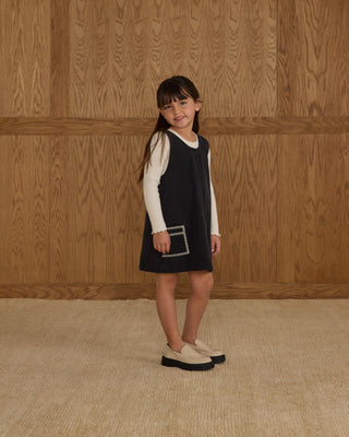 Rylee & Cru Ribbed Long Sleeve - Ivory
Young girl wearing a black dress with a white shirt underneath, standing against a wooden paneled wall.
