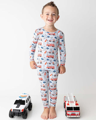 Lev Baby Pajama Set - Ryan
Child wearing pajamas with fire truck pattern, standing between two toy trucks on a white background