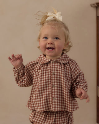 Rylee & Cru Bow Clip Set - Ivory
A smiling toddler with blonde hair tied up with a cream bow, wearing a brown and white gingham long-sleeve button-up shirt and matching gingham pants, standing against a plain beige wall.